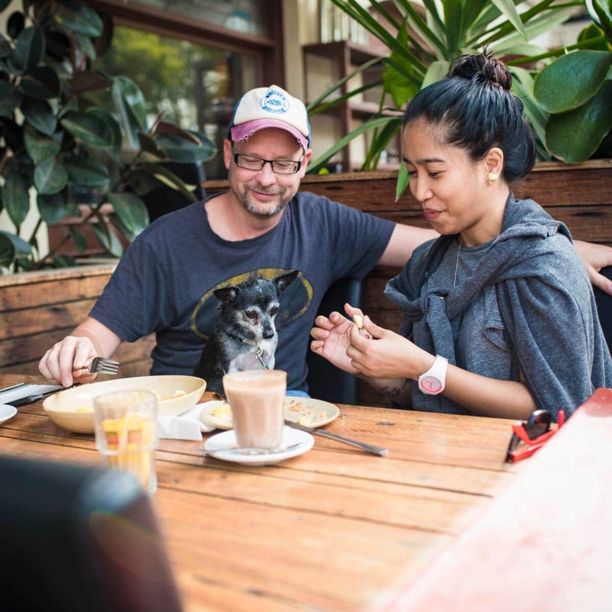 Two smiling individuals seated at a table in a dog cafe in Melbourne, with plates of food and drinks in front of them. They appear to be engaged in a cheerful conversation.