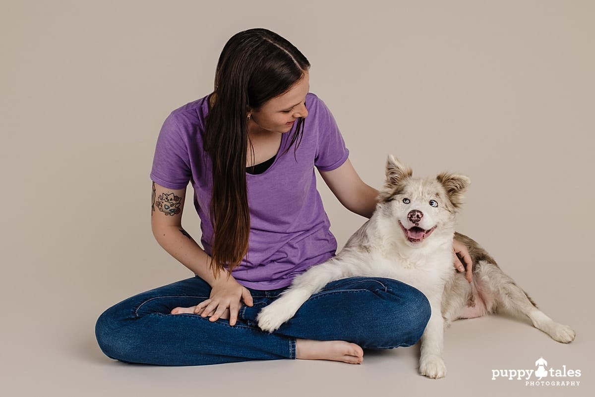 Dog photo of Merry, the Red Merle Border Collie with her person Charlotte. She’s wearing a purple shirt and blue jeans. They are posing right next to each other on a beige background for Project Dogalogue of Puppy Tales Photography.